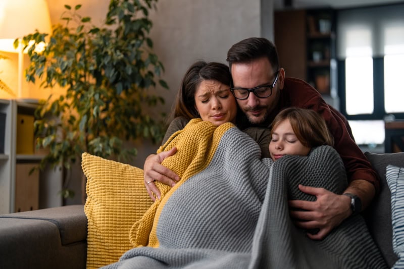 Married couple with child, bundled up on couch due to furnace not heating entire Wasilla, AK, home.